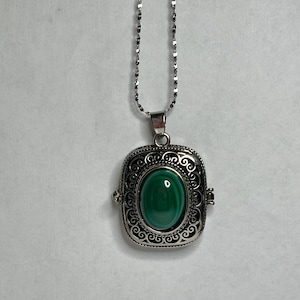 May include: A silver chain necklace with a square locket pendant featuring an oval-shaped green stone. The locket has an intricate, ornate design.