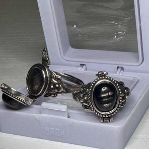 May include: Two silver-toned metal poison rings featuring oval-shaped black stones. One ring is shown with its hinged lid open, revealing a hollow compartment, while the other ring is closed, displaying the smooth, dark, polished stone surface.