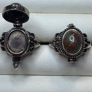 May include: Two silver-toned metal rings with oval settings. The ring on the left is open, revealing a hollow, textured interior compartment. The ring on the right is closed, featuring a smooth, multi-colored oval stone with brown and green tones.