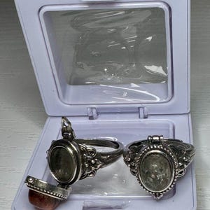 May include: Two silver-toned metal rings with oval-shaped locket compartments. One ring is open, revealing a hollow interior and a separate reddish-brown stone piece, while the other ring is closed, displaying a textured, light-colored oval stone.