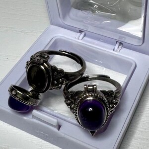 May include: Two silver-toned metal poison rings with oval settings. One ring is closed, featuring a smooth, deep purple cabochon stone. The other ring is open, revealing a hollow compartment, with its purple stone lid detached to the side.