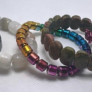 May include: A collection of beaded bracelets in various styles and colors. One bracelet is dark gray with a chevron pattern. Other bracelets feature white, rainbow, brown, blue, and star-shaped beads. The bracelets are displayed on a white surface.