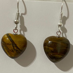 May include: A pair of heart-shaped earrings. Each earring features a polished, golden-brown stone with dark brown streaks. The stones are suspended from silver-colored fish hook ear wires. The earrings are displayed against a white background.