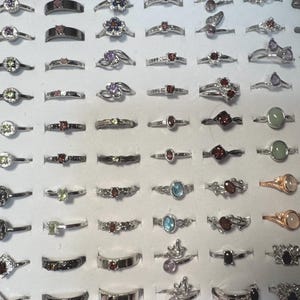 May include: A collection of various rings displayed in a white tray. The rings feature different designs, including gemstones in various colors such as red, green, and blue. The rings are made of silver-colored metal.