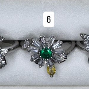 May include: Three silver rings with different designs. Ring 5 features a crescent moon and star design with clear stones. Ring 6 has a flower design with a green center stone and clear accents. Ring 7 showcases a butterfly design with clear stones.