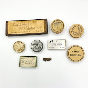 Op de afbeelding: Assortiment vintage horlogemakersbenodigdheden in ronde metalen en kartonnen dozen. De dozen zijn gelabeld met tekst in het Nederlands en Engels, waaronder "Cylinders volgens Sirius maat", "1 Gros Korkringe", "30 Marquisefedern" en "Bouchons pour Pendules".