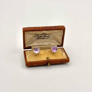 May include: A pair of gold-toned cufflinks with round, light purple centers and a ring of small, clear stones. The cufflinks are displayed in an open, wooden box with a tan velvet interior. The box has text that reads "10 Bold Street, Liverpool."