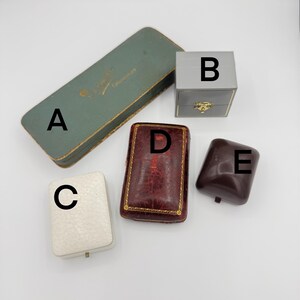May include: Five different empty jewelry boxes. The boxes are made of various materials, including cardboard, leather, and plastic. The boxes are in different colors, including green, gray, white, and brown. The boxes are labeled with letters A through E.