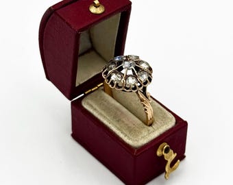 May include: A gold ring with a cluster of clear gemstones, set in a black and gold design, displayed in an open red jewelry box with a gold clasp. The ring's band has a floral design.
