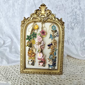 Puede incluir: Una colección de pendientes colgantes coloridos exhibidos en un marco dorado. Los pendientes presentan varios diseños, incluyendo flores, mariposas y casas. Los pendientes son en tonos de rosa, azul, amarillo y verde, con detalles metálicos dorados.