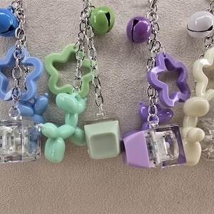 Puede incluir: Primer plano de varios colgantes coloridos colgando de cadenas plateadas. Los colgantes incluyen campanas azules, formas de estrella y figuras de animales de globo en varios colores pastel. Cada colgante está conectado a un pequeño objeto rectangular transparente.