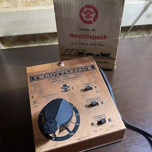 May include: A brown and copper colored model train throttle control with a black knob and a red overload indicator light. The control has a power switch, direction switch, and a dial that goes from 0 to 100. The text on the control reads "Throttlepack Model 501" and "Model Rectifier Corporation Edison, N.J. 14817".