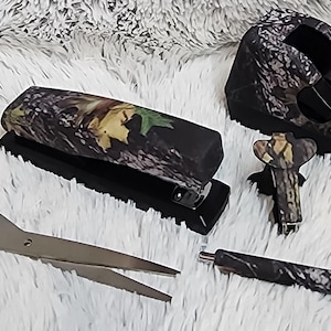 May include: A set of four camouflage-patterned office supplies: a stapler, a tape dispenser, a pair of scissors, and a pen. The items are all black with a green, brown, and tan camouflage pattern.