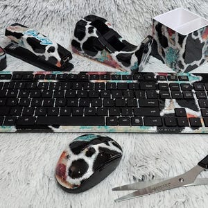 May include: A desk set featuring a keyboard, mouse, tape dispenser, stapler, pen holder, and scissors. The set has a black and white cow print design with accents of teal, red, and orange. The keyboard has a standard QWERTY layout.