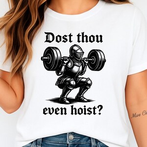 Pu&ograve; includere: T-shirt bianca con un'illustrazione in bianco e nero di un cavaliere che solleva un bilanciere. Il testo "Dost thou even hoist?" &egrave; stampato sopra e sotto l'immagine del cavaliere. Il design &egrave; centrato sulla maglietta.