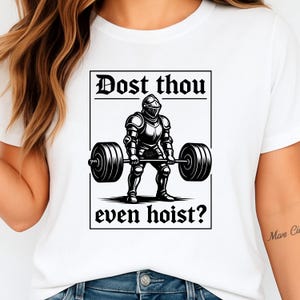 May include: White t-shirt featuring a black and white illustration of a knight lifting a barbell. The text "Dost thou even hoist?" is printed above and below the knight image. The design is enclosed in a black rectangular frame.