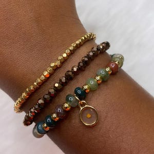 May include: Three gold beaded bracelets stacked on a wrist. The top bracelet is made of small gold beads, the middle bracelet is made of brown beads, and the bottom bracelet is made of green, brown, and red beads. A small gold charm with a clear glass center hangs from the bottom bracelet.