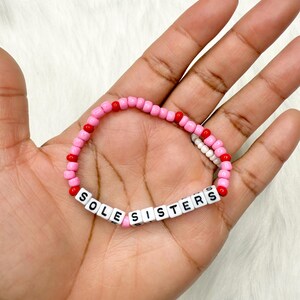 May include: A pink beaded bracelet with the words "SOLE SISTERS" spelled out in white letter beads. The bracelet features red and white accent beads. The bracelet is held in a hand.