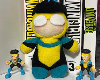May include: A plush doll of a character with black hair, white eyes, and a yellow, black, and teal suit, flanked by two small plastic action figures of the same character, all positioned in front of three books titled Invincible.
