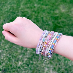Peut inclure: Un ensemble de bracelets en perles colorées sur un poignet. Les bracelets sont faits de petites perles rondes dans des tons de rose, violet, bleu, blanc et jaune.