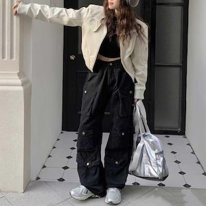 Puede incluir: Chaqueta color crema de corte corto, combinada con pantalones cargo negros con múltiples bolsillos y detalles plateados. El conjunto se completa con zapatillas plateadas y un bolso tote plateado. El look es casual y moderno.