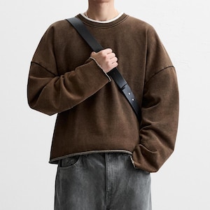 Puede incluir: Una sudadera marrón de manga larga con cuello redondo, dobladillo y puños sin rematar, sobre una camiseta blanca. La sudadera presenta hombros caídos y un corte holgado, combinada con unos pantalones vaqueros de mezclilla gris carbón.