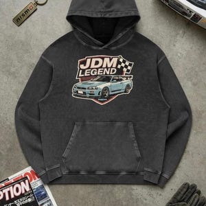 Può includere: Una felpa con cappuccio grigio antracite con una stampa grafica sul petto. Il disegno raffigura un'auto sportiva azzurra, una bandiera a scacchi e la scritta JDM LEGEND. La felpa è dotata di maniche lunghe, cappuccio e una tasca a marsupio.