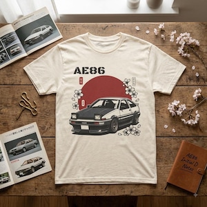 Könnte beinhalten: Ein cremefarbenes Kurzarm-T-Shirt mit einer Grafik eines schwarz-weißen AE86-Autos vor einem großen roten Kreis. Das Design enthält weiße Kirschblüten-Illustrationen und den in Schwarz über dem Auto gedruckten Text AE86.
