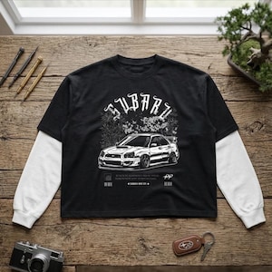 May include: A black long-sleeve t-shirt with white layered sleeves. The front features a white graphic illustration of a Subaru car, floral elements, and the text SUBARU in a stylised font, along with small text and barcode graphics below the car.