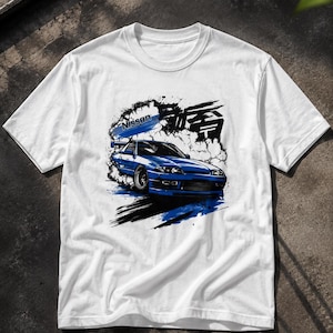 Op de afbeelding: Een wit t-shirt met korte mouwen en een grafische print van een blauwe Nissan sportwagen. Het ontwerp bevat zwarte en blauwe verfspatten, rookwolken en Japanse kanji-tekens naast het woord Nissan in blauwe letters.