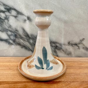May include: Ceramic candle holder with a beige base and a flared design. The holder features blue and brown leaf-like patterns and a circular base. The top and base rims are a darker beige color. The candle holder is on a wooden surface.