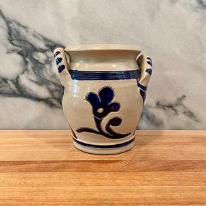 May include: A beige ceramic crock featuring two side handles and a hand-painted dark blue floral motif on the front. The vessel has a wide mouth, a cylindrical body, and horizontal blue bands encircling the top and bottom sections of the exterior.
