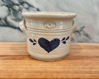 Może przedstawiać: Beżowy ceramiczny garnek z dwoma bocznymi uchwytami, ozdobiony namalowanym ciemnoniebieskim sercem z przodu oraz okrągłym wytłoczonym logo przy krawędzi z napisem Pinewood Valley. Naczynie posiada nakrapianą fakturę oraz szeroki, rozszerzający się otwór.