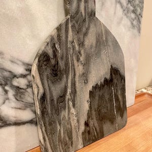 May include: A gray and white marble cutting board with a rounded top and a small hole for hanging. The stone features prominent dark gray and black veining patterns throughout the surface, creating a natural, swirling, and marbled appearance.
