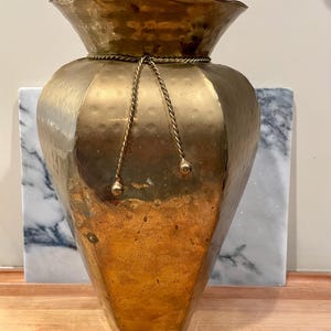 May include: A tall, hammered brass vase with a wide, scalloped rim and a tapered, multi-faceted body. A decorative twisted brass rope cord is tied around the neck, featuring two small, rounded finials hanging down the front surface.