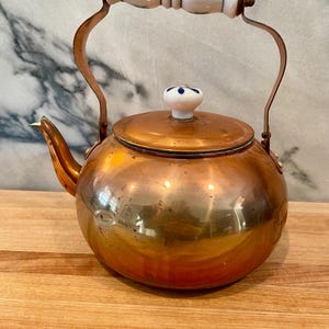 May include: A copper tea kettle with a white and blue floral patterned handle and lid knob. The kettle has a rounded body and a curved spout. The handle is made of ceramic and metal. The kettle sits on a wooden surface.