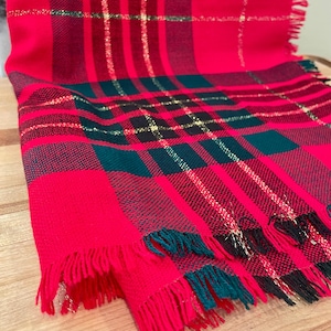 May include: A rectangular scarf featuring a vibrant red, dark green, and black plaid pattern. The fabric is woven with thin, shimmering gold metallic threads running through the design. The edges of the scarf are finished with a short, frayed fringe.