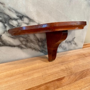May include: A wall-mounted shelf featuring a smooth, semi-circular wooden surface with a polished brown finish. The shelf is supported by a single, curved wooden bracket attached to the underside, providing a stable base for display.