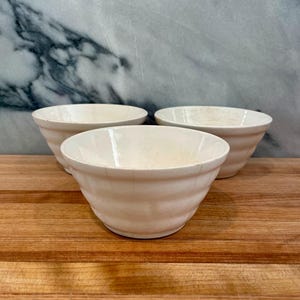 May include: Three off-white ceramic bowls with a ribbed design. The bowls are arranged on a wooden surface, with a marble background. The bowls are of a similar size and shape, suitable for serving food.