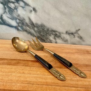 May include: A brass spoon and a three-tined brass fork featuring black handles with decorative metal accents at the base. The utensils have a polished gold-tone finish on the bowl and tines, with a smooth transition to the dark handle sections.
