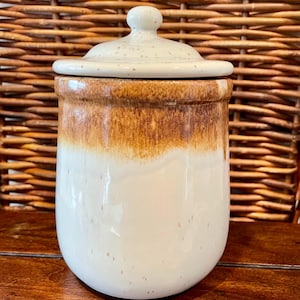 May include: A ceramic jar with a lid. The jar is white with a brown ombre effect around the top. The lid is off-white with small brown speckles. The jar is sitting on a wooden surface.