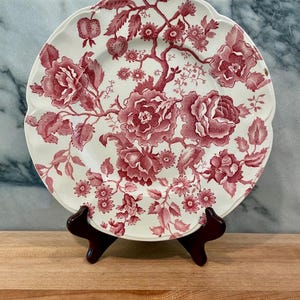May include: A round ceramic plate with a scalloped edge, featuring a detailed red floral pattern on a white background. The design includes various blooming flowers, leaves, and small fruit-like elements spread across the surface.