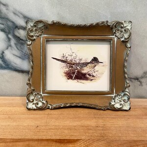 May include: A rectangular picture frame with an ornate, distressed bronze and white finish containing a print of a roadrunner bird standing in a desert landscape with sparse vegetation and a light-colored background.