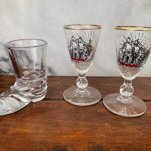 May include: Three clear glass shot glasses. One is shaped like a boot, and the other two are stemmed glasses with a gold rim and a black, red, and white ship design. The glasses are on a wooden surface.