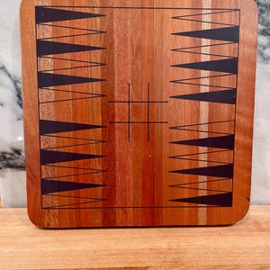 May include: A square wooden backgammon board featuring a natural reddish-brown wood grain finish. The board displays two sets of twelve black triangular points arranged on opposite sides, with a central black grid pattern etched into the surface.