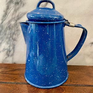 May include: A blue enamelware coffee pot with a lid and handle. The pot has a speckled white pattern and a spout for pouring. The handle is attached with a metal hinge. The pot is sitting on a wooden surface.