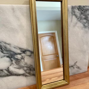 May include: A rectangular wall mirror featuring a slim, metallic gold-toned frame with a beveled edge. The reflective glass surface is clear and smooth, capturing a view of an interior space including a wooden door in the background.