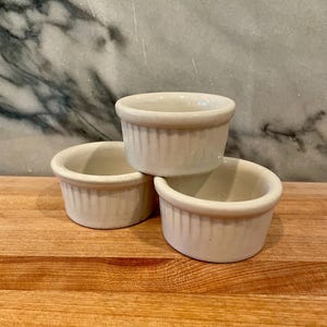 May include: Three small, off-white ceramic ramekins stacked on a wooden surface. Each ramekin has a fluted exterior and a rounded rim. The background features a marble-patterned surface.