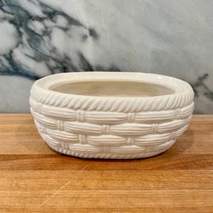 May include: An oval-shaped white ceramic planter featuring a textured exterior that mimics the appearance of a woven basket. The vessel has a smooth, glossy finish and a wide, open top, providing a clean and neutral aesthetic for holding plants.