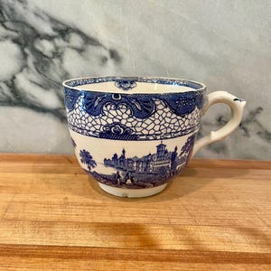 May include: A white ceramic teacup featuring a blue transferware pattern. The upper rim displays a decorative lattice design, while the lower section depicts a detailed landscape scene with buildings and figures. It has a curved handle.